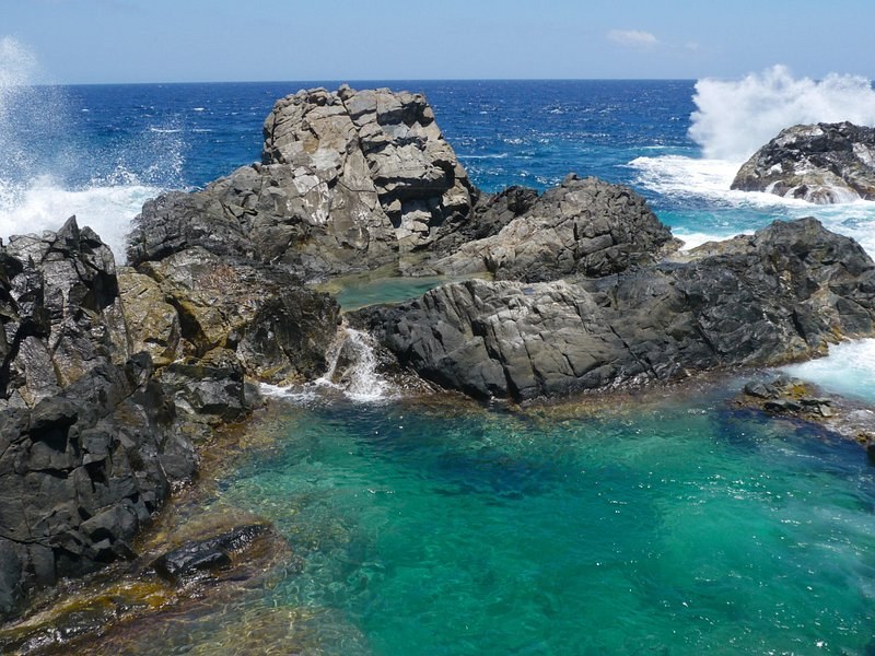 Surf breaking over black rock on the windward coast, with a clear turquoise tidal pool held behind it