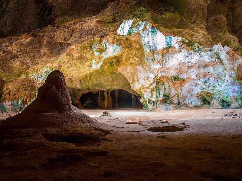 A cave chamber in the park, its walls stained pale green and ochre by mineral seepage, with a sand floor and a rock mound at the entrance